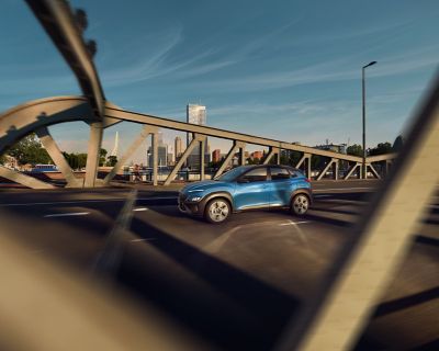 The new Hyundai Kona from the front in Surfy Blue driving through a busy city street.