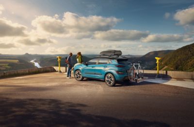 The Hyundai Kona Electric with the accessories tow bar, bike carrier and roof box on a parking lot.