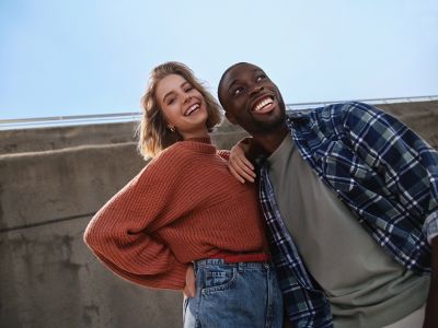 Eine junge Frau in rotem Pullover und ein junger Mann in kariertem Hemd lachen Arm in Arm.