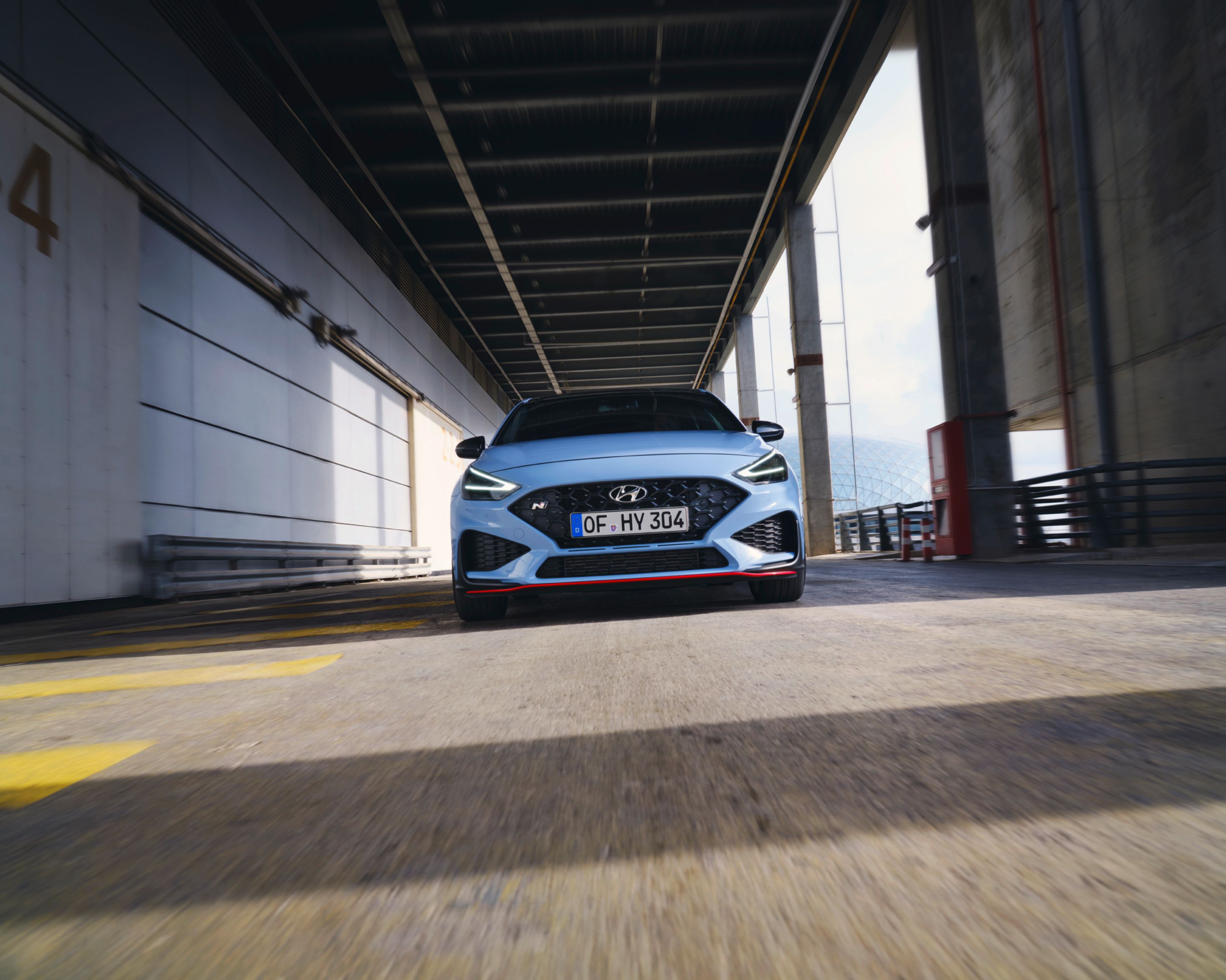 The new Hyundai i30 N from the front in Performance Blue colour driving along an industrial structure.