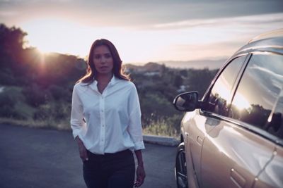 Eine Frau in weißer Bluse steht neben einem Hyundai Fahrzeug. Im Hintergrund eine hügelige Landschaft mit Sonnenuntergang.