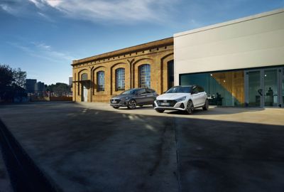 Nuova Hyundai i20 N Line e i20 in versione Aurora Grey parcheggiate accanto a un grande edificio.