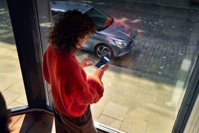 Ragazza guarda fuori dalla finestra la sua Hyundai i20 con uno smartphone in mano con app Bluelink aperta