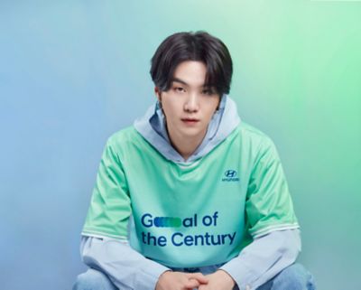 BTS member Suga wearing a green Hyundai Team Century shirt with a gray hoodie.