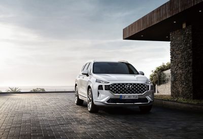 The new Santa Fe 7 seat SUV in white from the front parked in front of a house with ocean view.