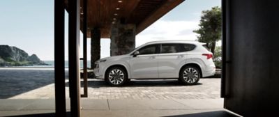 The Santa Fe 7 seat SUV in white from the side parked in front of a house with ocean view.