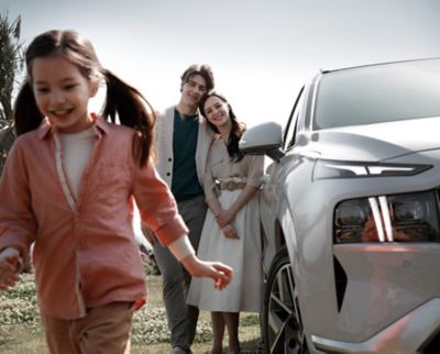 Ein Mann und eine Frau stehen lächelnd aneinander gelehnt neben einem weißen Hyundai SANTA FE. Ein Mädchen mit 2 braunen Zöpfen läuft lachend vor ihnen.