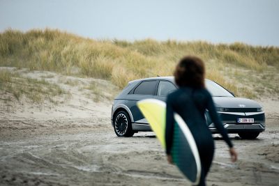 Kobieta z deską surfingową idzie w kierunku swojego samochodu IONIQ 5 zaparkowanego na plaży.