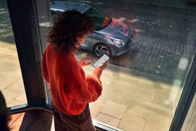 A woman looking at the Hyundai Bluelink App on her smartphone, her Hyundai i20 is parked outside.