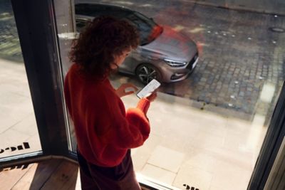 Eine Frau steht mit Smartphone am Fenster und blickt auf ihren draußen geparkten Hyundai i30..