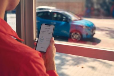 Kobieta stoi przy oknie z telefonem i łączy się za pomocą aplikacji Bluelink ze swoim samochodem.