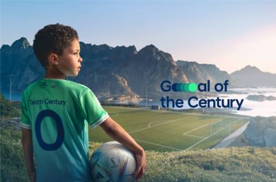 Chłopiec z piłką stoi odwrócony plecami na tle boiska piłkarskiego z logo Goal of the Century.