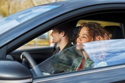 Eine junge Frau am Steuer eines Hyundai mit halb offenem Fenster. Daneben ein junger Mann.