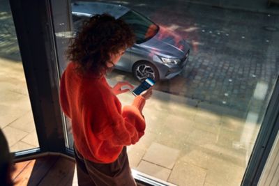 Una joven que mira su smartphone detrás de una gran ventana con un nuevo i20 aparcado fuera.