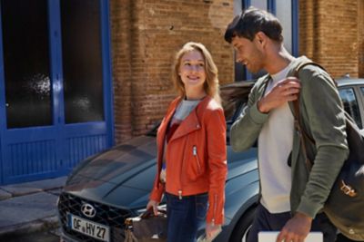 Eine junge Frau in roter Jacke und ein junger Mann gehen an einem geparkten Hyundai i20 vorbei.