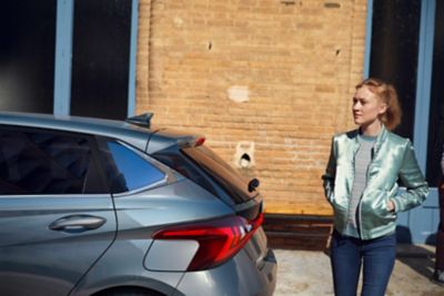 Mladá žena stojaca za novým modelom Hyundai i20, pohľad z ľavej zadnej strany