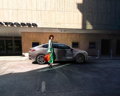 Fashionable woman in a green dress and with a red shopping bag walking towards her Hyundai i30 Fastback