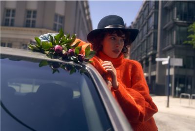 Kobieta w pomarańczowym swetrze i kapeluszu trzymająca kwiaty opiera się o samochód Hyundai i30.