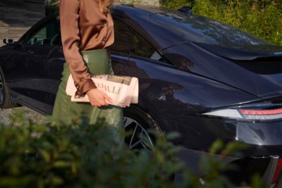 Woman with brown t-shirt and green skirt holding a newspaper walk next to the blue Hyundai IONIQ 6