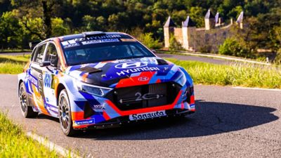 Hyundai i20 N Rally2 racing on a mountain road with a castle in the background.