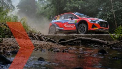 Hyundai Motorsport customer racing a Hyundai i20 N Rally2 car on forest road near a stream.