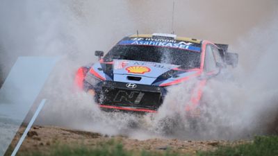 The Hyundai i20 N WRC rally race car speeding through a large puddle.