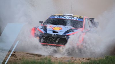 The Hyundai i20 N WRC rally race car speeding through a large puddle.