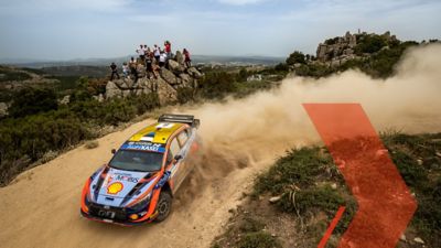 The Hyundai i30 N WRC rally race car taking a sharp turn on a dirt road.