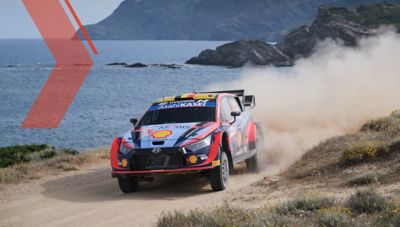 The Hyundai i20 N WRC rally race car taking a sharp turn on a dirt road next to the ocean.