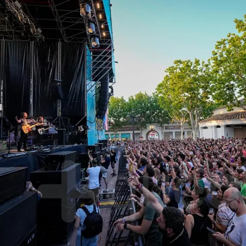 Vista lateral de un concierto al atardecer con escenario, árboles y público levantando los brazos.