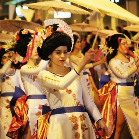 Mujeres con vestidos blancos y sombrillas bailando en desfile nocturno.