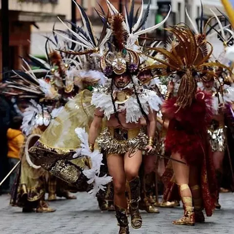 Comparsa con trajes dorados y grandes tocados de plumas avanzando por una calle adoquinada.