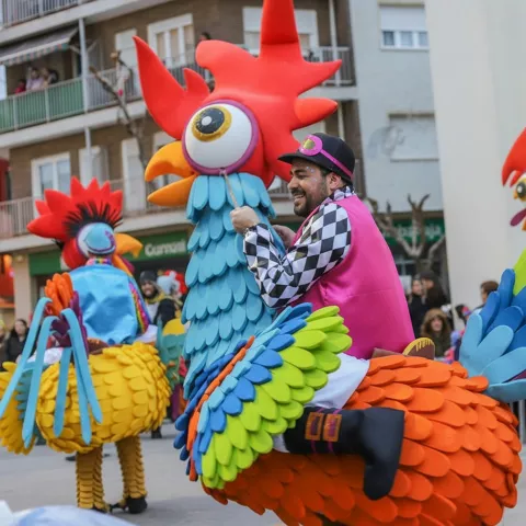 Participantes montados en grandes figuras de gallos multicolores durante desfile.