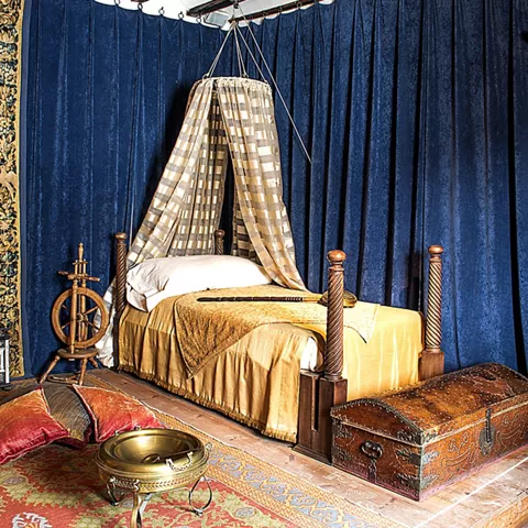 Dormitorio histórico con cama con dosel, baúl decorado y rueca de madera.