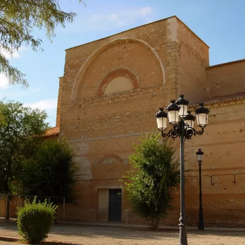 Muro de iglesia de ladrillo y piedra con arco decorativo, árboles y farolas delante.
