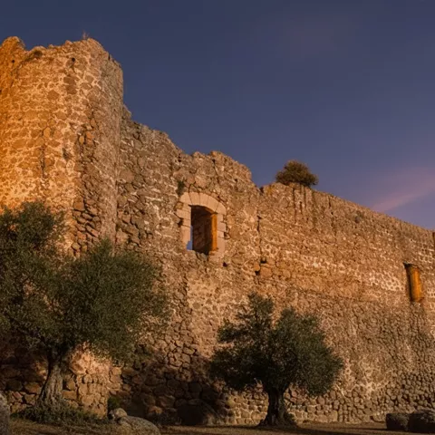 Ruinas de castillo de piedra al atardecer con torres circulares y olivos alrededor.