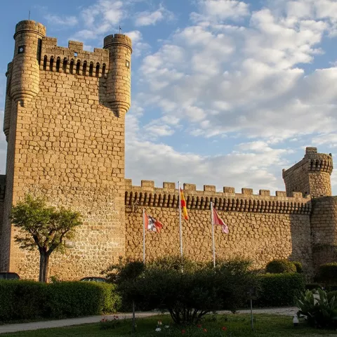 Castillo de piedra con torres almenadas y banderas frente a jardín.