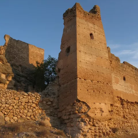 Torre y muros de piedra y tapial en un cerro rocoso, iluminados por el sol.
