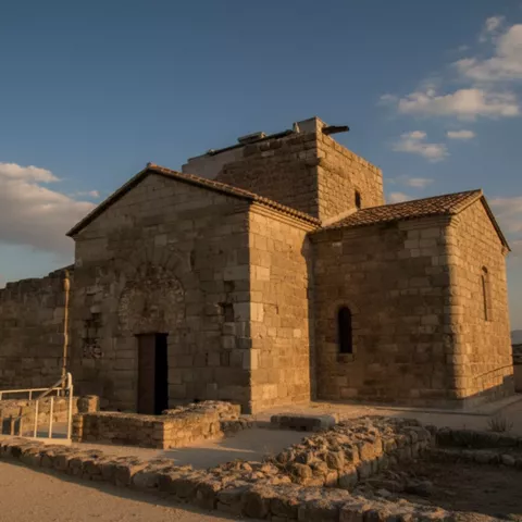 Ermita de piedra con ábside y torre, iluminada por luz cálida al atardecer.