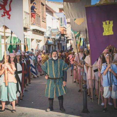 Desfile histórico por una calle con participantes portando estandartes y escudos.