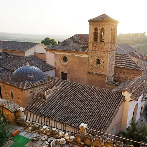 Vista aérea de iglesia de ladrillo con torre campanario y cúpula, rodeada de tejados antiguos al atardecer.