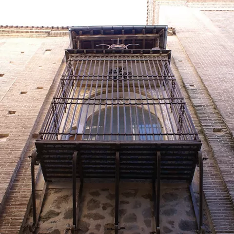 Balcón enrejado sobre fachada de ladrillo y piedra visto desde abajo.