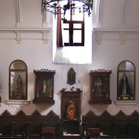 Capilla con pinturas y esculturas religiosas sobre muros blancos y sillería de madera.