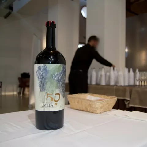 Botella de vino sobre mesa con mantel blanco, con copas y servicio al fondo desenfocado.