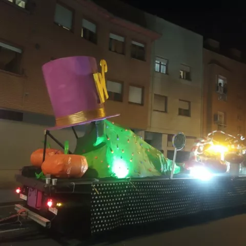 Carroza con sombrero gigante y figuras iluminadas avanzando por avenida urbana.