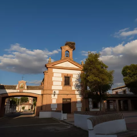 Conjunto urbano tradicional, con torre campanario, arco de paso y bancos en una plaza soleada.