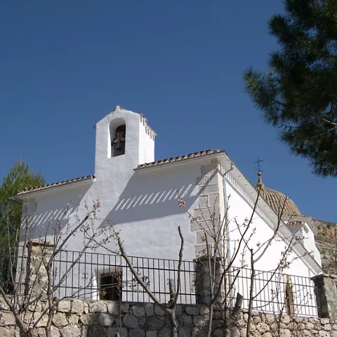 Vista trasera de ermita blanca con espadaña y cúpula entre árboles y colina.