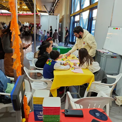 Taller infantil con un adulto ayudando a varios niños con piezas de colores en una mesa amarilla.