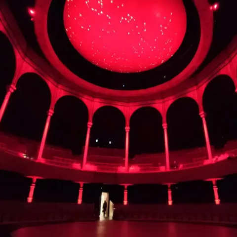 Interior circular iluminado en rojo con arcos y cúpula decorada.