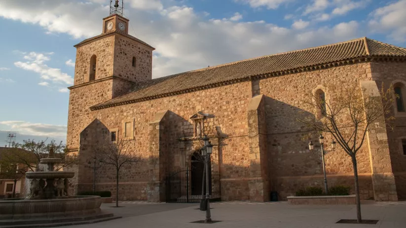 Iglesia parroquial de Fernán Caballero (Ciudad Real) con torre campanario de piedra y plaza con fuente en primer plano.